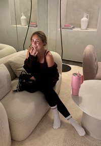 Borsa nera su un divano di peluche di colore chiaro; un tumbler rosa con una cannuccia su un tavolo bianco; tessuti morbidi e texture; abbigliamento casual e calzini bianchi.