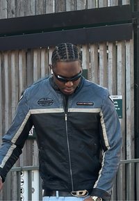 Veste de moto en cuir noir et bleu avec des rayures argentées, fermeture éclair à l'avant et écussons logo, portée avec des lunettes de soleil et des écouteurs.