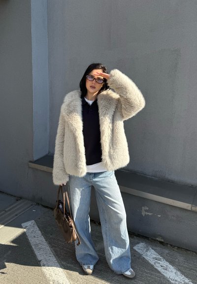 Chaqueta de pelo sintético en crema, corte holgado, con un amplio cuello. Combinada con una blusa oscura y unos pantalones vaqueros de pierna ancha en azul claro, sosteniendo un bolso marrón.