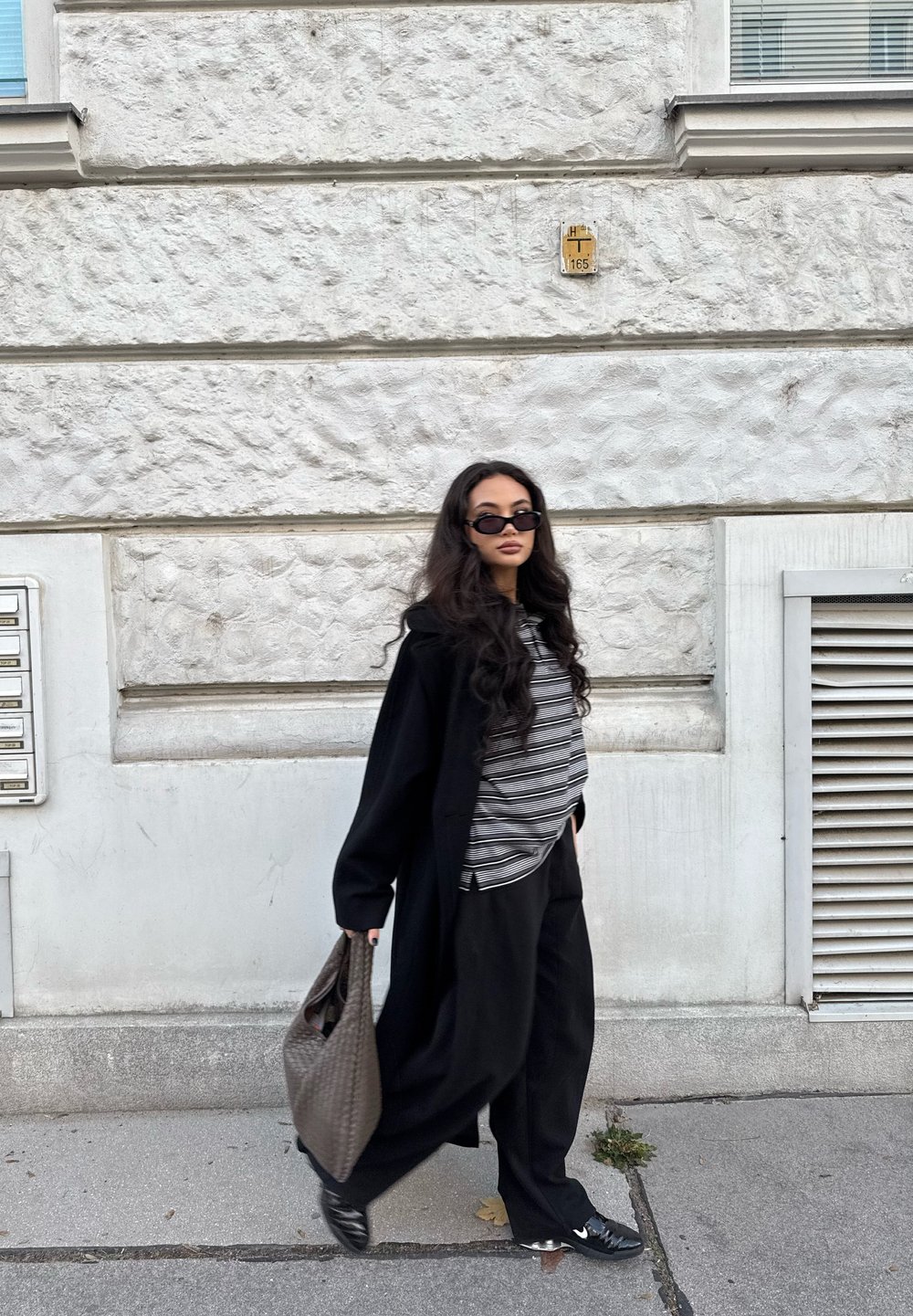 Casaco longo preto sobre uma blusa riscada, calças largas pretas e óculos de sol pretos. A pessoa carrega uma bolsa cinza trançada e está encostada numa parede texturizada.
