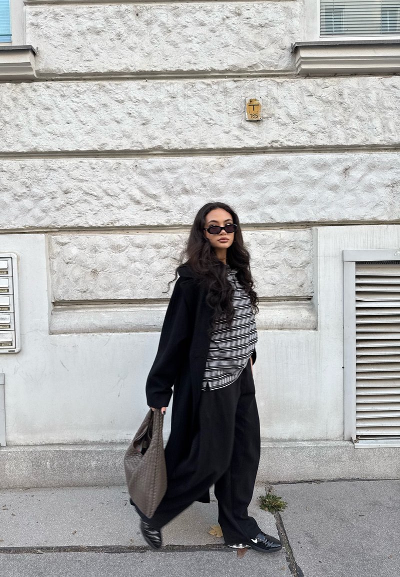 Palton lung negru peste un top cu dungi, pantaloni negri largi și ochelari de soare negri. Persoana poartă o geantă țesută de culoare gri și stă sprijinită de un perete texturat.