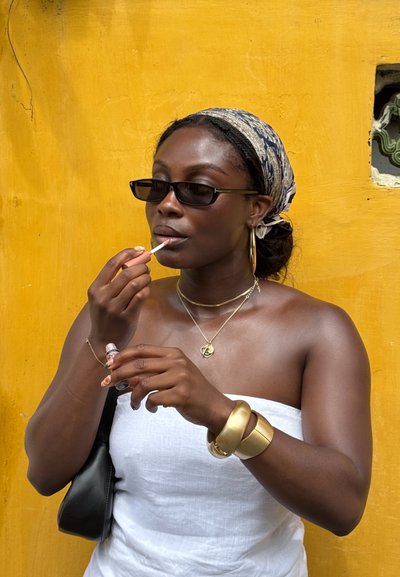 Gafas de sol negras, joyas de oro y un pañuelo de cabeza estampado con cabello largo. Aplicándose brillo labial mientras lleva una blusa blanca sin tirantes frente a una pared amarilla.