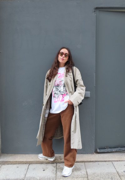 Abrigo trench beige con forro de cuadros sobre una camiseta gráfica blanca y pantalones de pierna ancha marrones. Fondo de pared gris, zapatillas blancas.