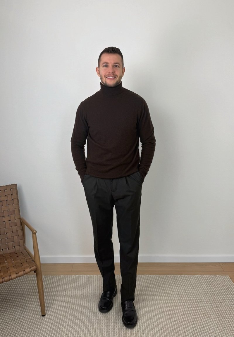 Man standing indoors wearing a dark brown turtleneck, black pants, and black shoes, with hands in pockets and smiling.