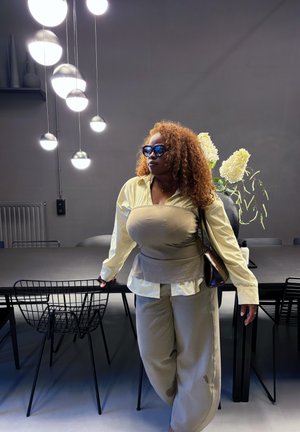 Chemise jaune clair superposée sur un haut beige, portant un pantalon beige assorti. Lunettes de soleil rondes noires, avec un sac à main texturé et un décor minimal en arrière-plan.