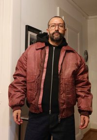 Ūdensizturīga tumši sarkana bombera jaka ar ribotām aprocēm, pārmērīgu fit, rāvējslēdzēju priekšpusē un divām priekšpusē novietotām kabatām, pārģērbta pāri melnam kapučjakai.