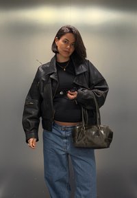 Veste noire en cuir oversize à texture côtelée, haut noir, jean taille haute bleu clair, et petit sac à main sombre.