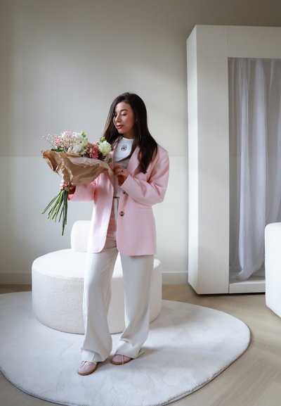 Blazer de gran tamaño en rosa claro sobre una blusa gris, pantalones blancos y zapatos en rosa suave. Sosteniendo un ramo con flores blancas y rosas, sobre una alfombra redonda.