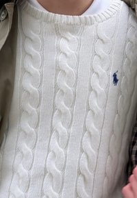 Pull blanc en maille torsadée avec logo Polo Ralph Lauren bleu brodé sur la poitrine, porté par-dessus une chemise blanche et une veste beige.