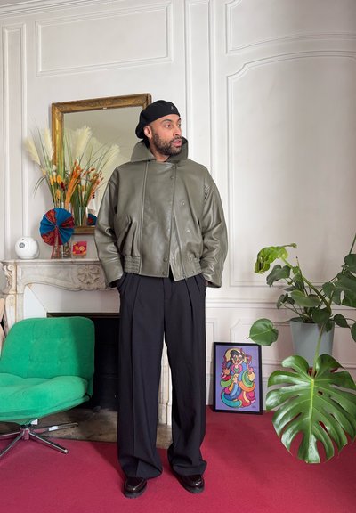Hombre con boina negra, chaqueta de cuero verde oliva y pantalones negros de pie en una habitación ornamentada con una silla verde, una planta grande y una obra de arte colorida.
