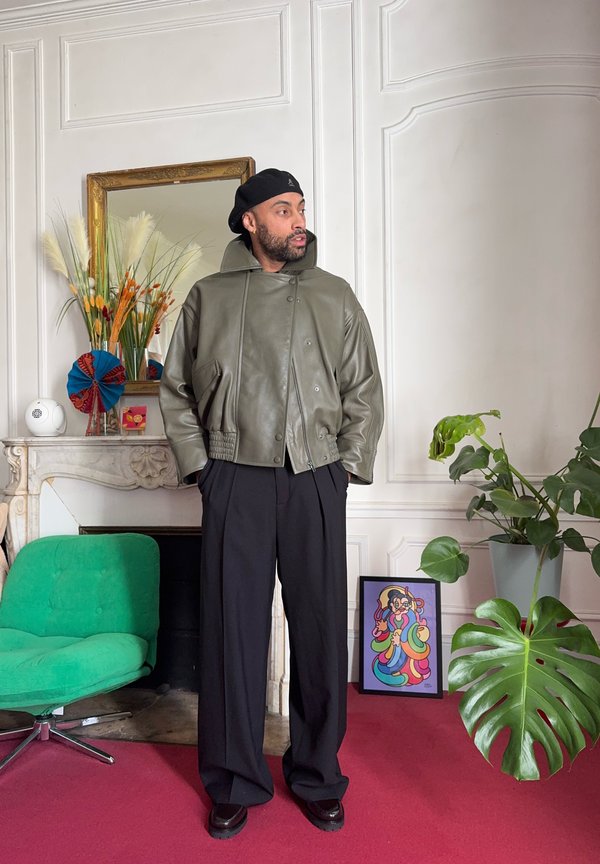 Vyras juodu baretu, alyvuogių žalios odinės striukės ir juodų kelnių stovi puošniame kambaryje su žalia kėde, dideliu augalu ir spalvingu meno kūriniu.