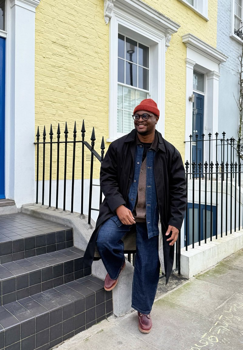 Manteau noir sur un jean en denim, pull marron, jeans à jambes larges, bonnet rouge en tricot et bottes marron. Assis sur des marches carrelées noires près d'un mur jaune.