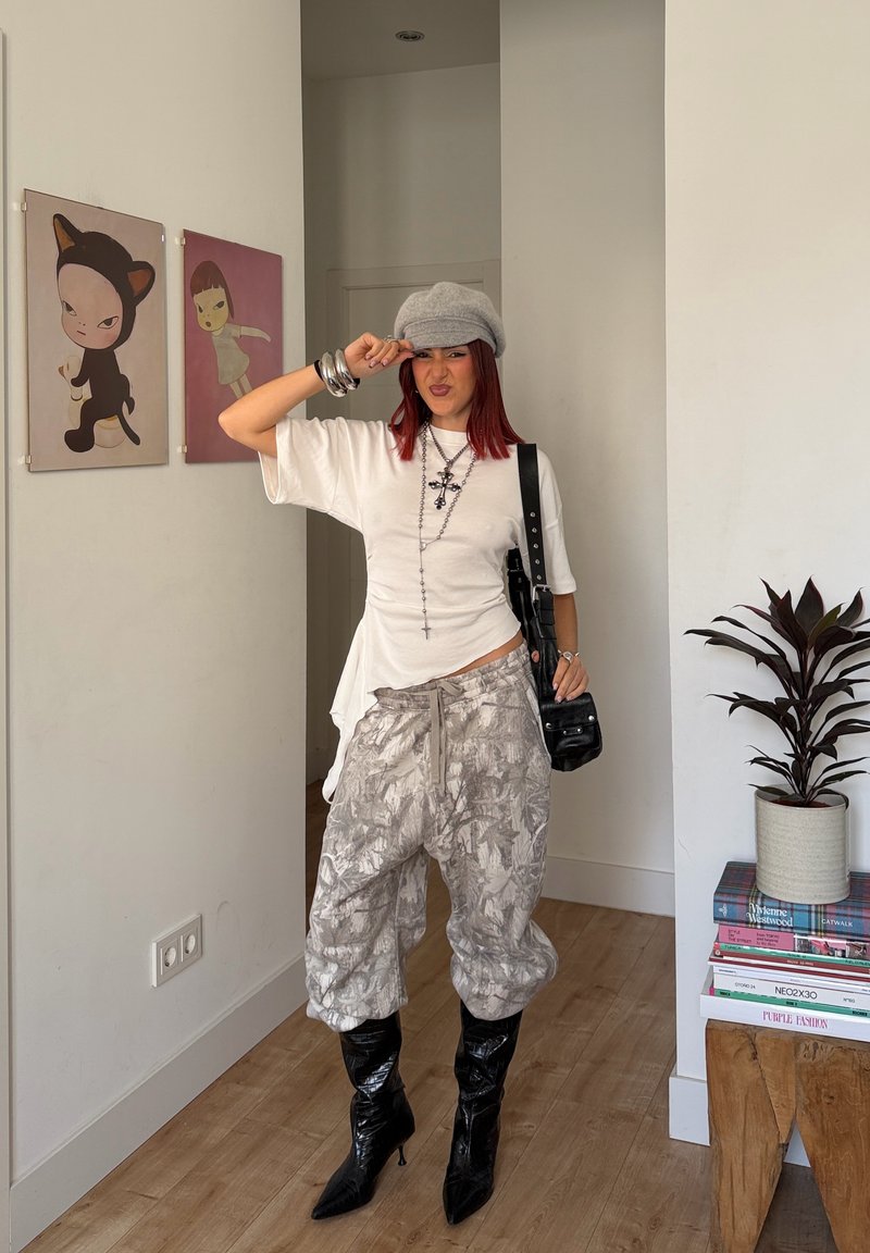 Young woman with red hair wearing a gray cap, white top, patterned baggy pants, and black heeled boots, posing indoors near artwork and plants.