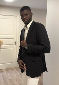 Schwarze maßgeschneiderte Blazer, über einem weißen Hemd getragen, mit Vorderentaschen und einer körperbetonten Passform. Lange Ärmel und eine Uhr am linken Handgelenk.
