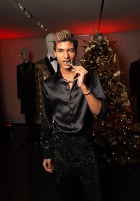 Jeune homme en chemise satin noire et pantalon scintillant mord un collier pendentif coloré lors d'un événement festif, avec un arbre de Noël doré et des mannequins en arrière-plan.