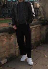 Casaco de couro castanho sobre camisola cinzenta, calças pretas largas e ténis brancos. Estar de pé junto a um corrimão de pedra num ambiente ao ar livre.