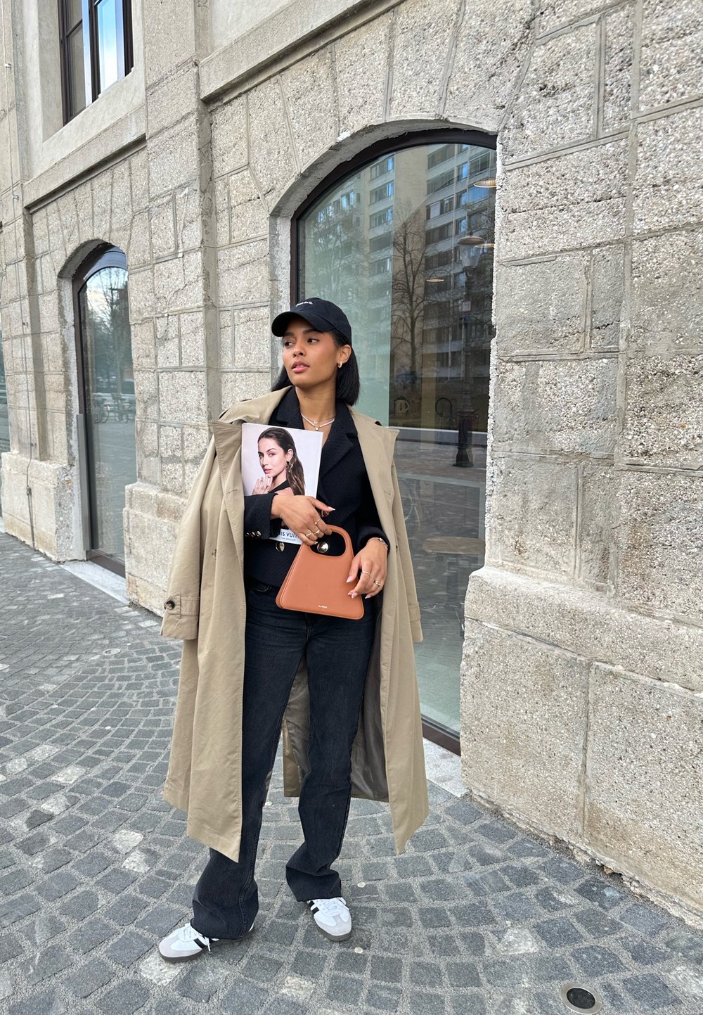 Beige trenchcoat över en svart outfit, håller en mörkbrun handväska och en tidning. Vita sneakers, står på en kullerstensgata.
