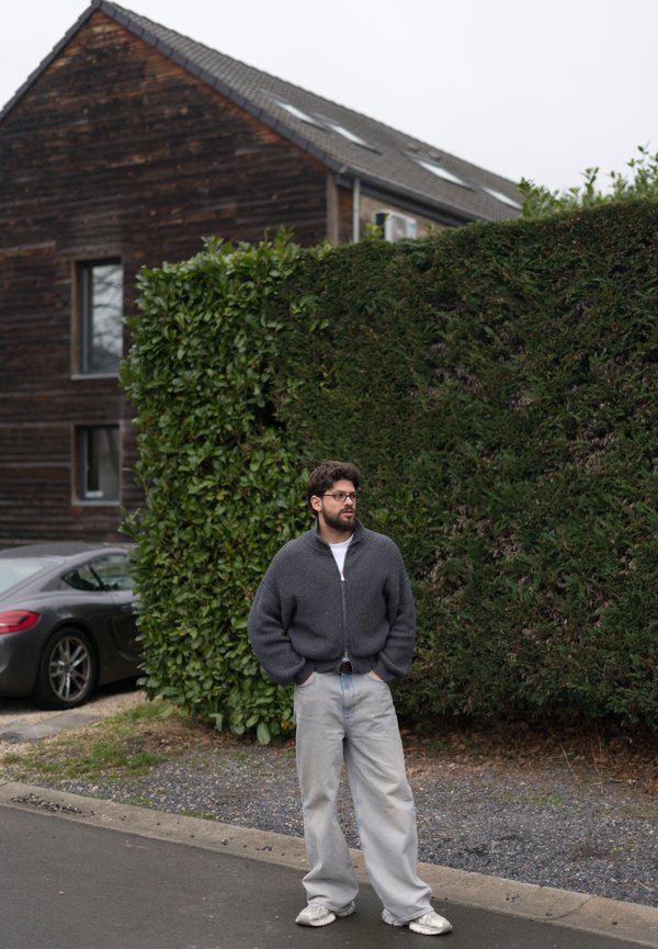 Pelēks džemperis ar rāvējslēdzēju, gaiši zilas plašas džinsu bikses un gaiši sporta apavi. Apkārt ir dzīvžogs un fonā redzama koka māja.