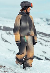 Άτομο με καφέ και γκρι εξοπλισμό σκι, μαύρο κράνος με πορτοκαλί γυαλιά σκι, στέκεται σε χιονισμένο βουνό, κοιτάζοντας προς το πλάι.