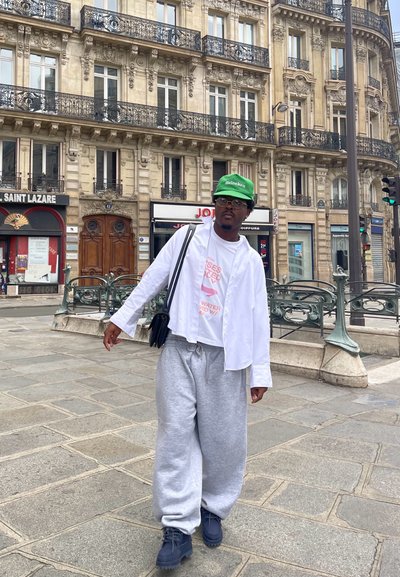 Conjunto casual que incluye una camisa blanca oversized, una camiseta gráfica rosa, pantalones de chándal grises y una gorra verde de Heineken. Un bolso negro y botas completan el look.