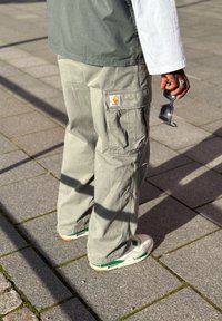 Jasnoszare spodnie cargo z wieloma kieszeniami i logo Carhartt. W zestawieniu z białymi i zielonymi trampkami, stojąc na kamiennej nawierzchni.