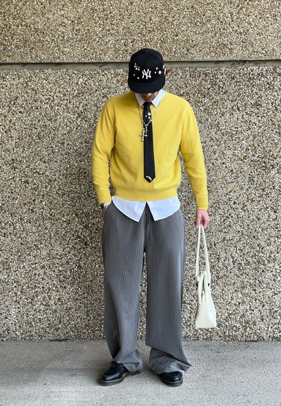 Suéter amarillo sobre una camisa blanca, corbata negra y pantalones de pierna ancha gris con rayas. Gorra negra con adornos; sostiene una bolsa blanca.