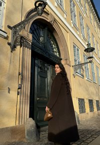 Καφέ παλτό από μαλλί, πράσινη διακοσμημένη πόρτα με καλλιτεχνική αψίδα, πέτρινη βάση και μπεζ τοίχο. Σκιές που ρίχνονται από το ηλιακό φως.