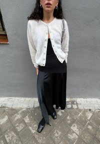 Heledane beež pehme kardigan, millel on nööbid, seljas must soonikkoes vormis top, kombineeritud musta satiinist maksiseeliku ja ruudukujuliste mustade kingadega.