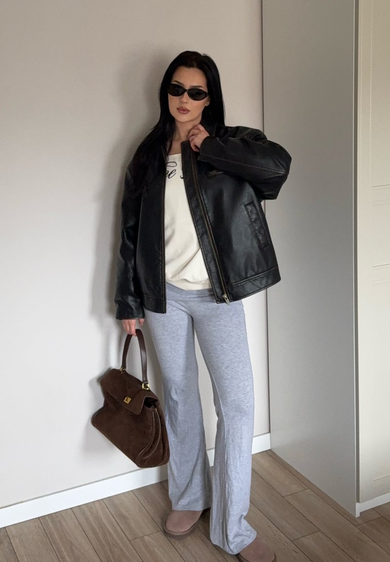 Femme portant une veste en cuir noire, un haut blanc, un pantalon gris, des lunettes de soleil, tenant un sac à main marron, debout à l'intérieur sur un sol en bois.