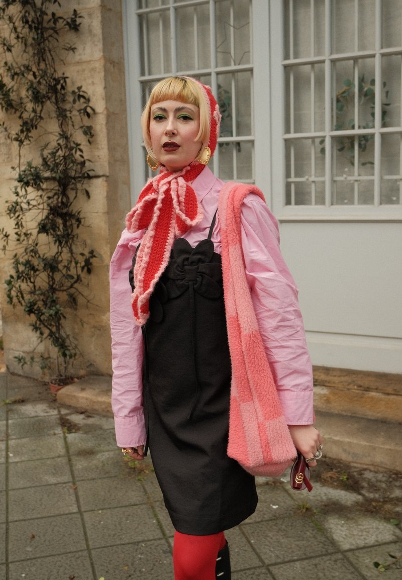Person mit kurzen blonden Haaren, trägt ein pinkes Hemd, ein schwarzes Kleid, rote Strumpfhosen, einen rot-pinken gestrickten Schal und eine Mütze, trägt eine große pinke Tasche.
