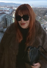 Manteau en fourrure brune, grandes lunettes de soleil noires, sac à main noir texturé avec détails dorés, debout devant un décor urbain.