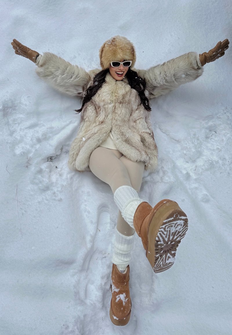 Femme en manteau de fourrure, chapeau et bottes allongée dans la neige, les bras étendus, faisant un ange de neige, souriante et portant des lunettes de soleil blanches.