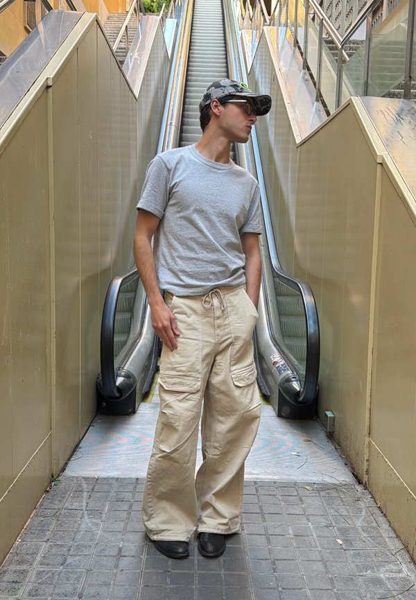 Junger Mann im grauen T-Shirt, beiger Cargohose und Kappe steht vor einer Außentreppe und schaut nach links.