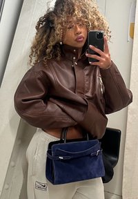 Personne aux cheveux bouclés portant une veste en cuir marron et un pantalon beige, tenant un sac à main en daim bleu marine, prenant un selfie dans un miroir avec un smartphone.