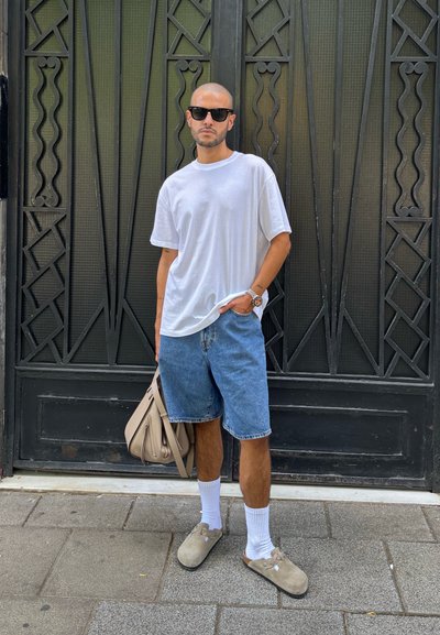 Camiseta blanca de algodón, pantalones cortos de mezclilla azul, zapatos grises sin cordones, calcetines blancos y bolso beige; de pie frente a una puerta decorativa negra.