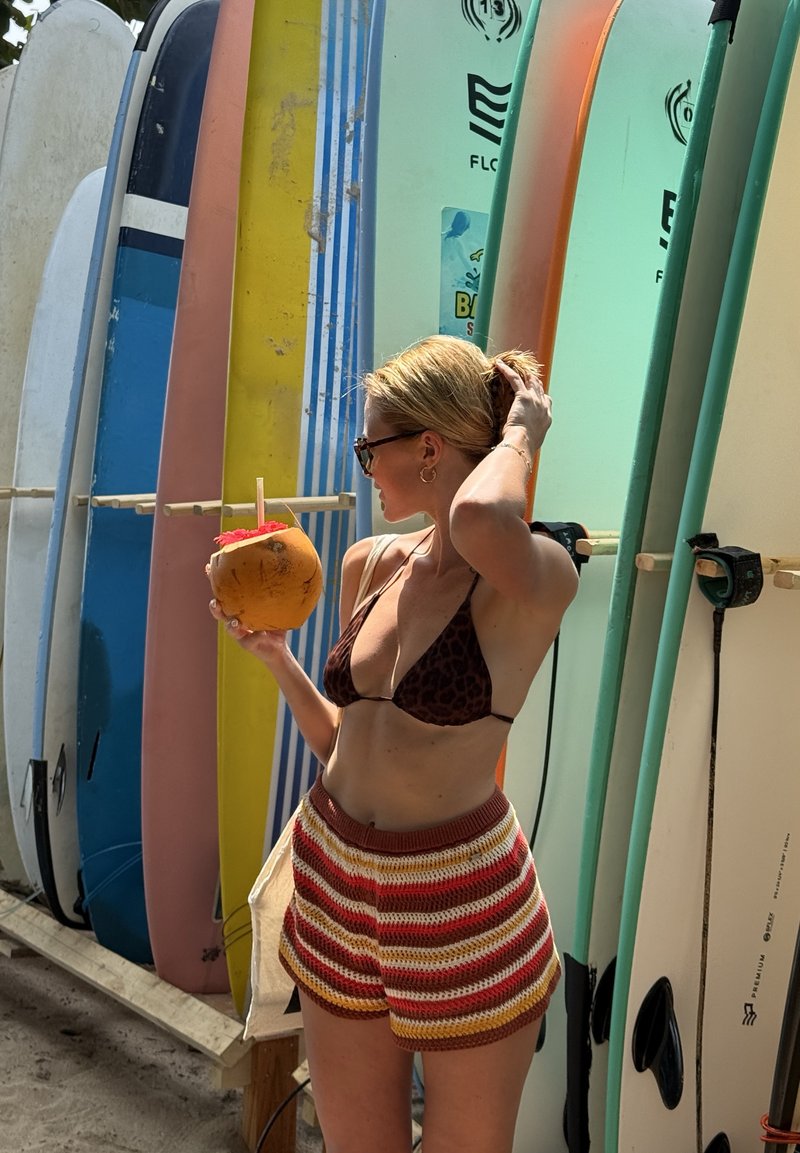 Donna con top bikini e pantaloncini a righe tiene una bevanda in cocco, in piedi accanto a un supporto con tavole da surf colorate sulla sabbia.