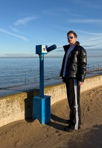 Homme en doudoune noire et pantalon Adidas se tient à côté d'un télescope bleu à pièces au bord de la mer par une journée ensoleillée.