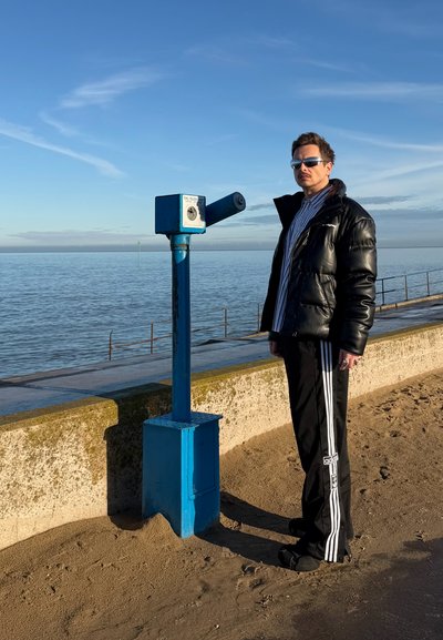 Hombre con chaqueta acolchada negra y pantalones Adidas junto a un telescopio azul de monedas junto al mar en un día soleado.