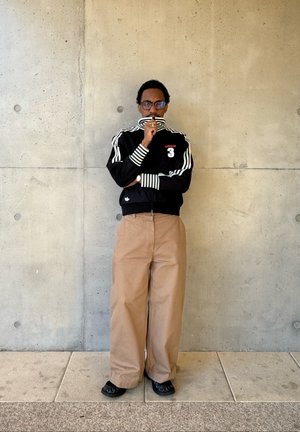Hombre con gafas que lleva una chaqueta deportiva negra de Adidas y pantalones anchos beige, de pie contra una pared de hormigón con los brazos cruzados.