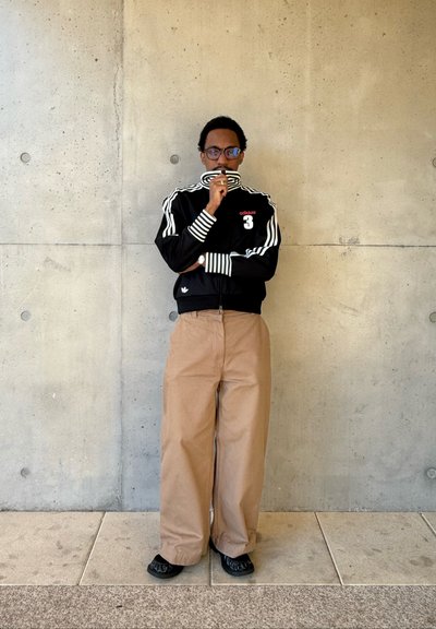 Hombre con gafas que lleva una chaqueta deportiva negra de Adidas y pantalones anchos beige, de pie contra una pared de hormigón con los brazos cruzados.