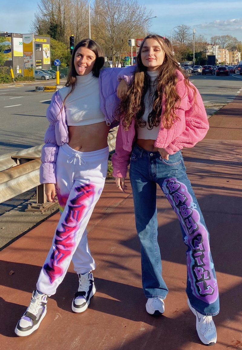 To kvinder står på en fortov, iført cropped tops og farverigt overtøj. Den ene har en lavendel farvet pufferjakke på, den anden i pink. Unikke grafiske bukser.