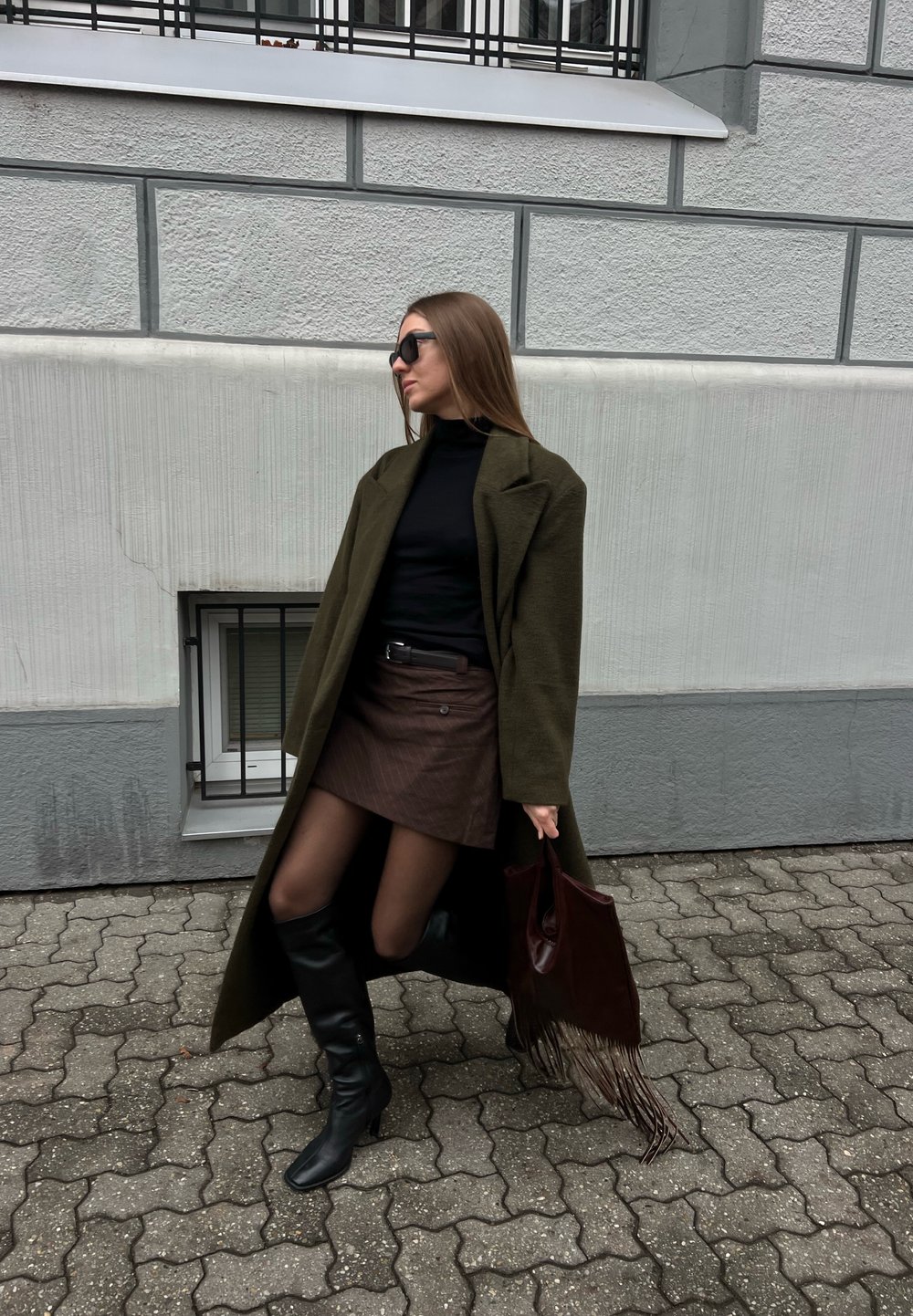 Femme portant des lunettes de soleil, un long manteau olive, un col roulé noir, une jupe marron, des bottes noires, tenant un sac à franges, debout sur un trottoir pavé.