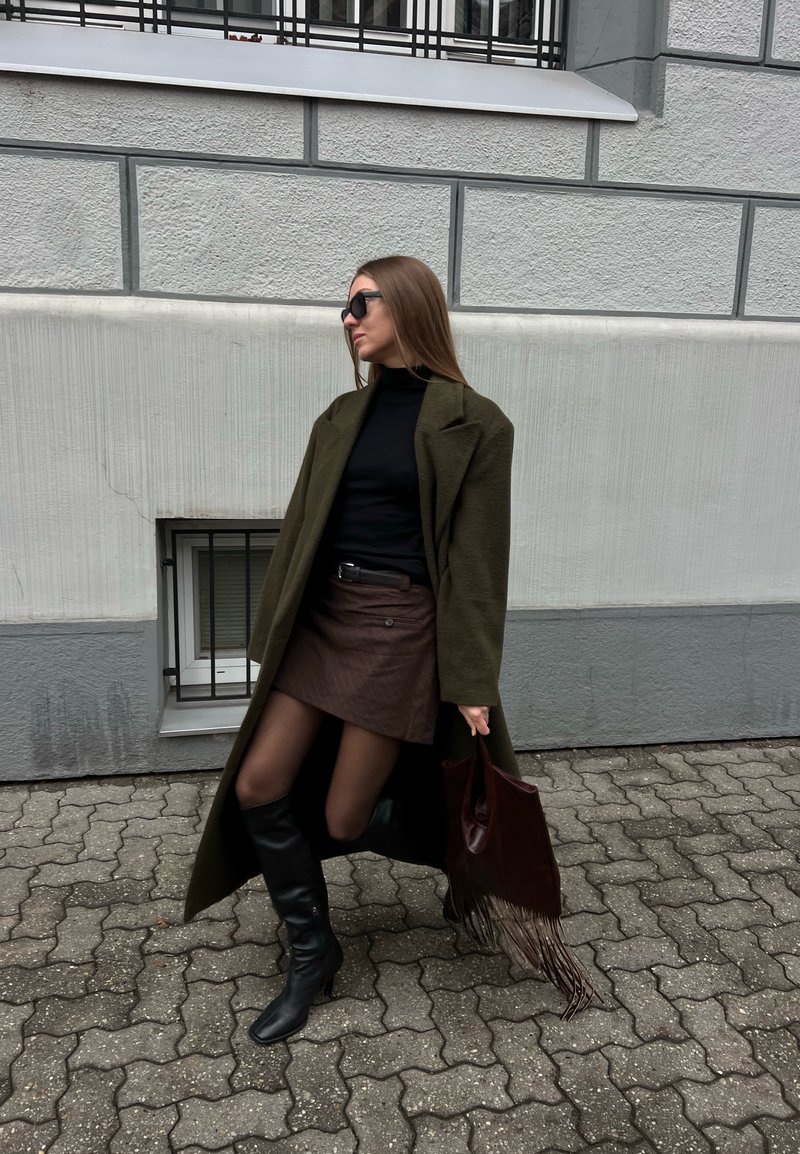 Woman in sunglasses wearing a long olive coat, black turtleneck, brown skirt, black boots, holding a fringe handbag, standing on a paved sidewalk.