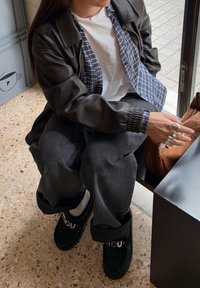 Personne assise à l'intérieur près d'une fenêtre, portant une veste en cuir noire, une chemise à carreaux blanche et bleue, un pantalon ample foncé, des chaussures noires, tendant la main vers un sac marron.