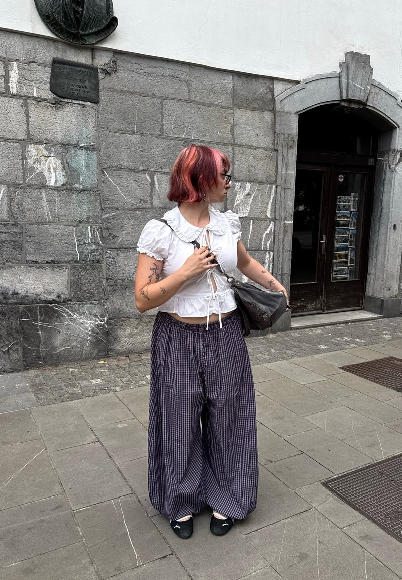 Jeune personne aux cheveux roses et tatouages se tient sur un trottoir en pierre près d'un bâtiment en briques grises et d'une porte en bois foncé.