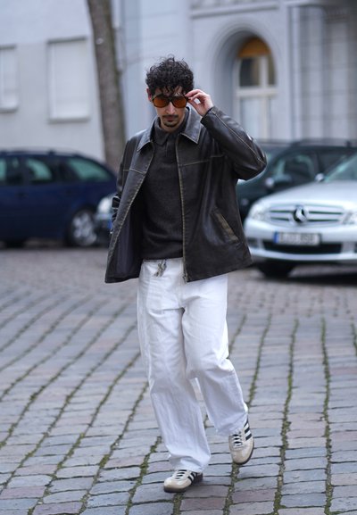 Chaqueta de cuero negra, suéter gris, pantalones blancos holgados y zapatillas con rayas negras. Gafas de sol marrones. Fondo de calle adoquinada.