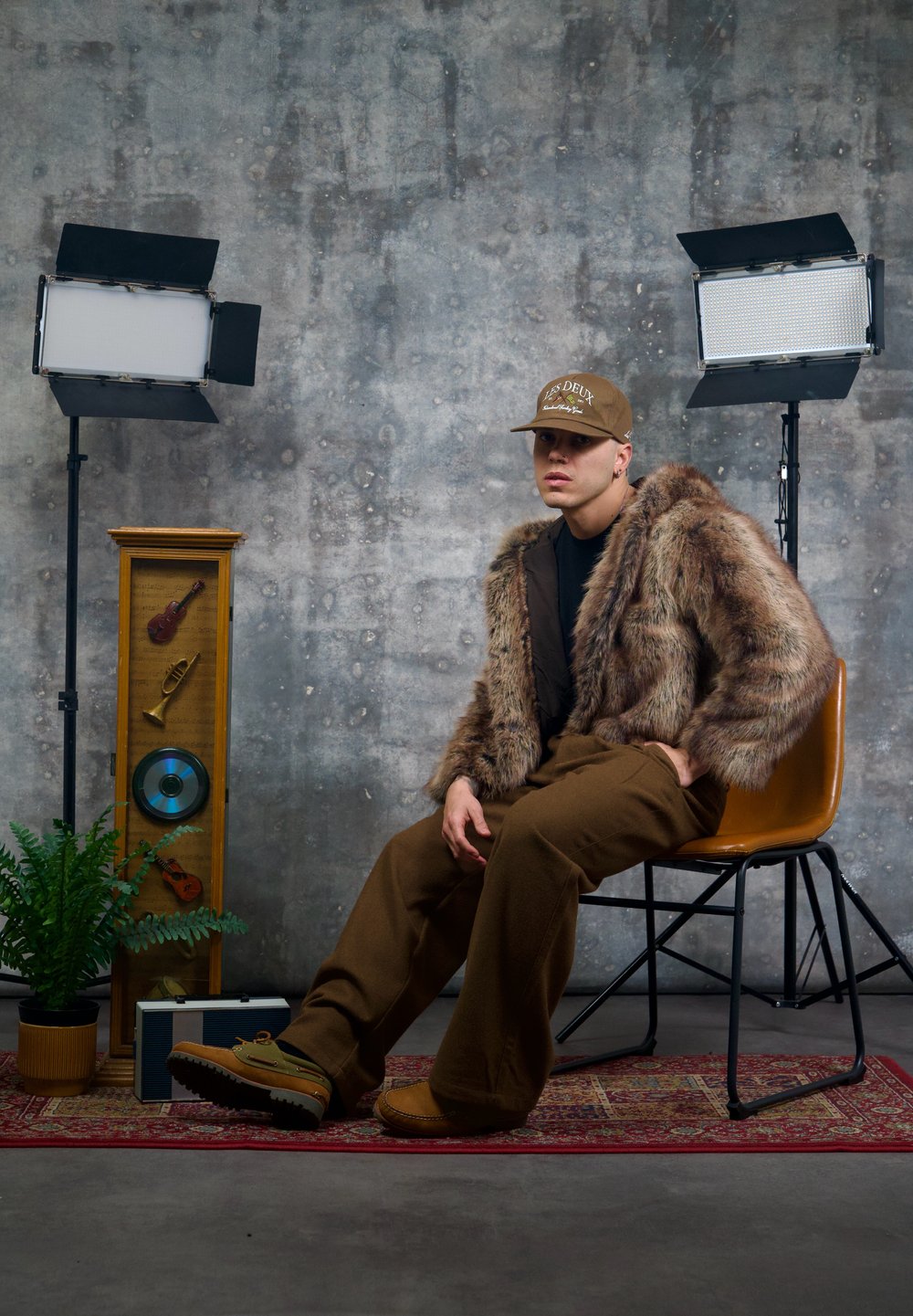 Veste en fausse fourrure marron, chemise noire, pantalon large marron, casquette marron, assis sur une chaise devant un décor comprenant des plantes et des éléments décoratifs.