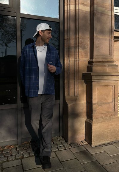 Camisa de cuadros azul sobre una camiseta blanca, pantalones grises y zapatillas negras. De pie cerca de un muro de piedra y una puerta de cristal, con una gorra blanca.