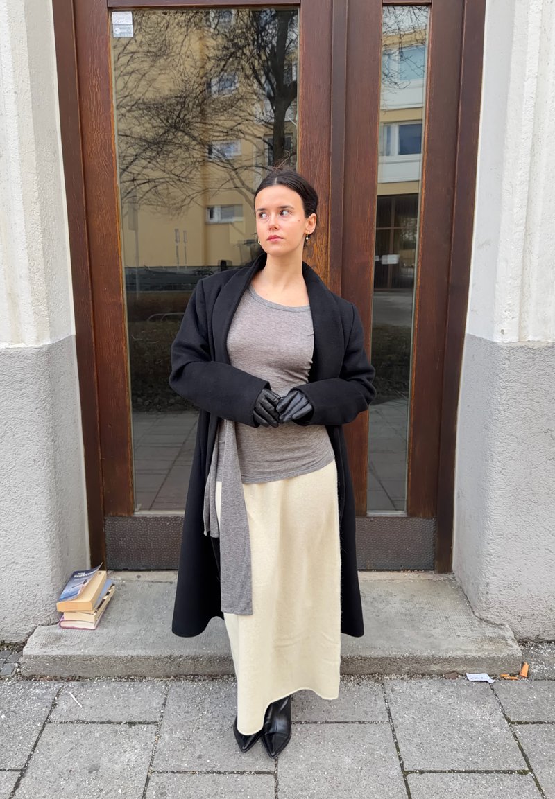 Femme portant un haut gris, une jupe crème, un manteau noir et des gants, se tient les mains jointes devant une porte en bois, des livres empilés sur le trottoir à proximité.