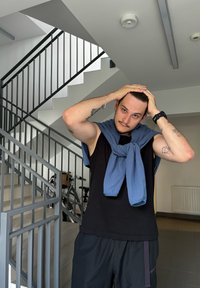 Fekete ujjatlan felső, fekete rövidnadrág, kék pulcsi a vállon. Beltéri környezet szürke lépcsőkkel és kerékpárokkal a háttérben.